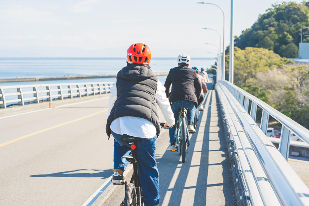 縦に連なりながら海沿いの道路をサイクリングするグループ